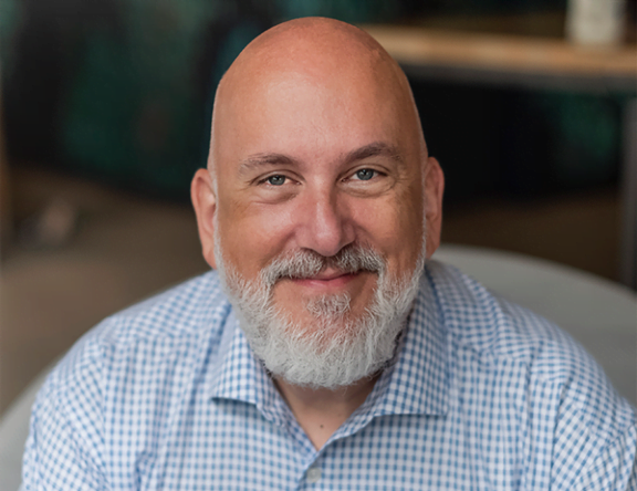 A picture of Mark Goad, smiling with blue eyes, white beard and a blue checkered shirt