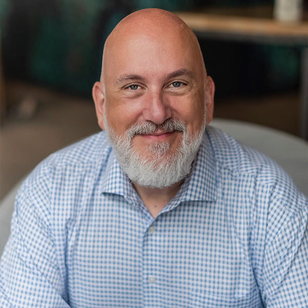 A picture of Mark Goad, smiling with blue eyes, white beard and a blue checkered shirt